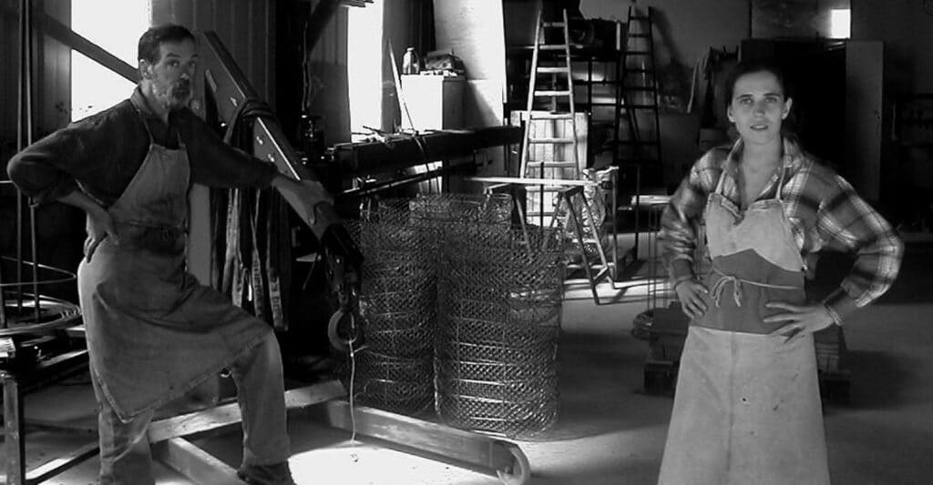 Deux ouvriers en tabliers posent dans un atelier métallique. Échelles, cages de grillage et matériel industriel en arrière-plan.