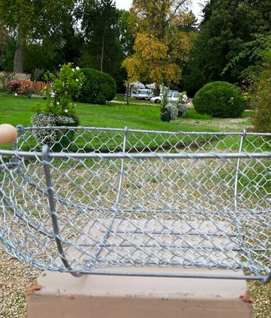 Panier vide en grillage métallique au premier plan, avec un jardin verdoyant, des arbres, buissons et véhicules stationnés en arrière-plan.